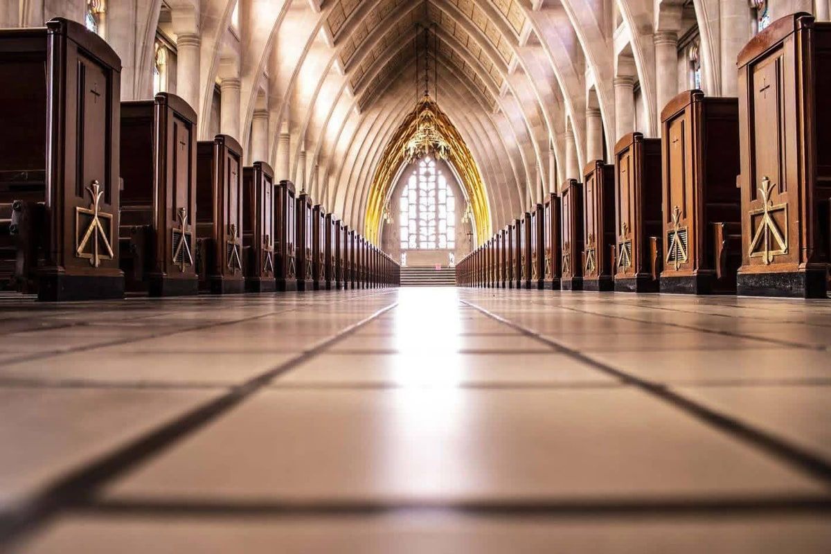 A church interior photographed from floor level: a long tiled aisle between two rows of dark wooden pews with small gold crosses, stone arches overhead, and a bright arched window glowing at the far end.