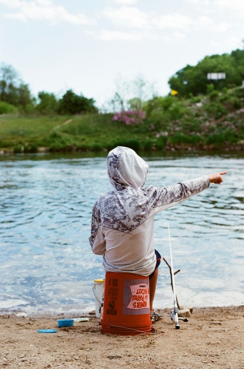 A child in a gray camouflage hoodie sitting on an overturned orange five-gallon bucket on a sandy riverbank, a fishing rod propped alongside, pointing across the water toward the far green bank.