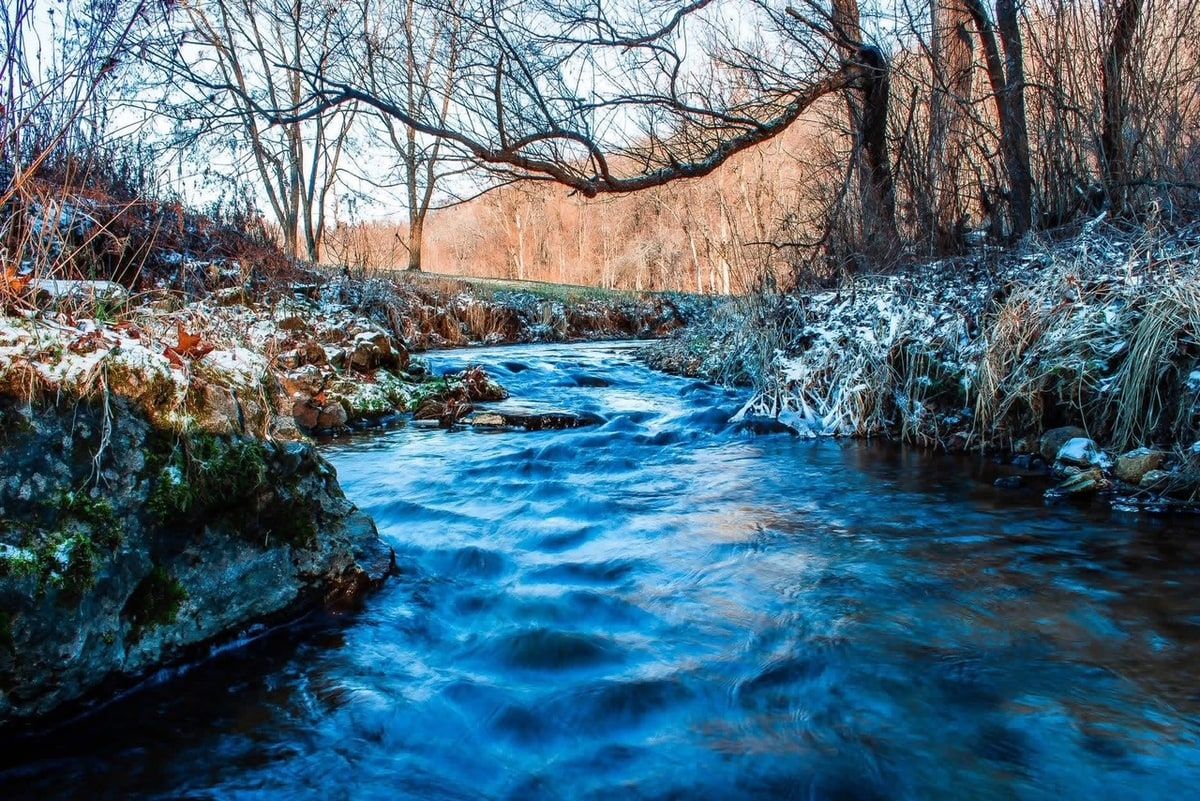A creek running open between frost-covered banks, the moving water blurred soft and blue by a long exposure, low sun on the far hillside.
