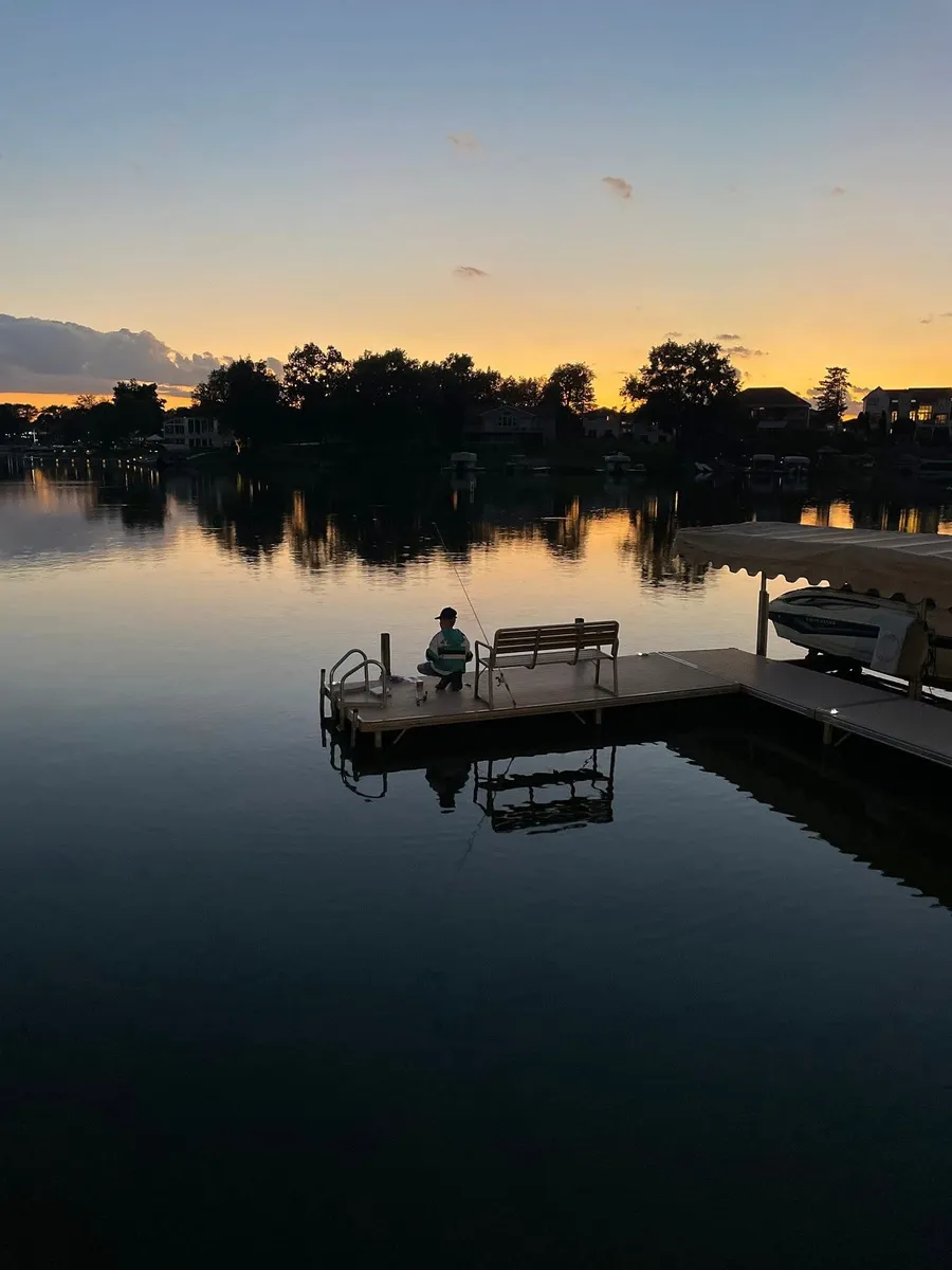 A child sits alone on a small dock at dusk, fishing rod cast out over calm water, cap and striped jersey silhouetted against the fading light. An empty bench stands behind them on the dock. Houses and trees line the far shore, their shapes and a few lit windows reflected in the still water, and a boat rests under a lift canopy at the dock’s near edge.