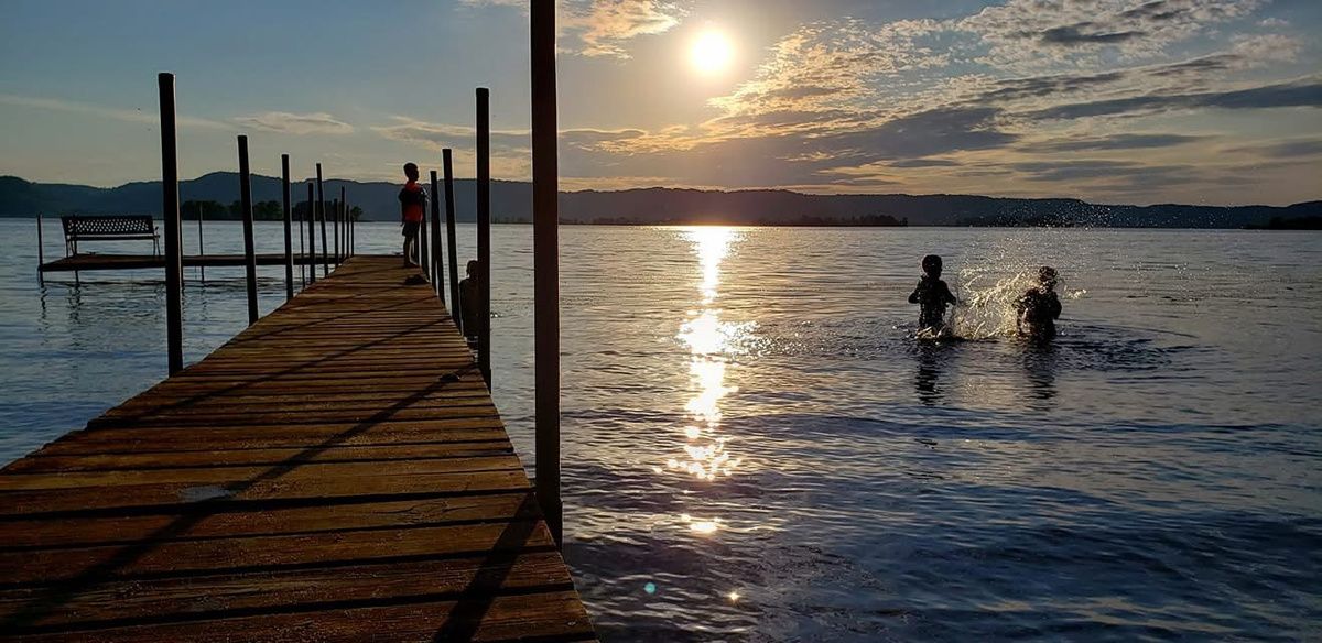 The same dock and lake at sunset, undarkened and carrying no type. A child stands partway down the dock in silhouette, another crouched near the water at its far end. Two more kids splash in the lake beyond the dock, backlit against the sun's reflection. Low hills line the far shore, and an empty bench sits on a side platform near the dock's near end.